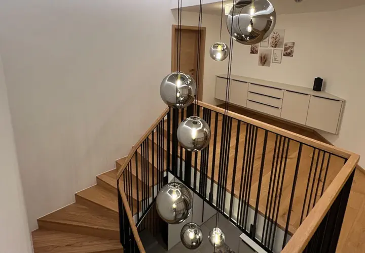 Nowoczesna klatka schodowa z drewnianymi stopniami i czarną balustradą, ozdobiona wiszącymi kulistymi lampami.