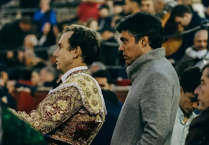 Un torero con traje de luces bordado y zapatillas torero, junto a un hombre con abrigo gris, observan la plaza.