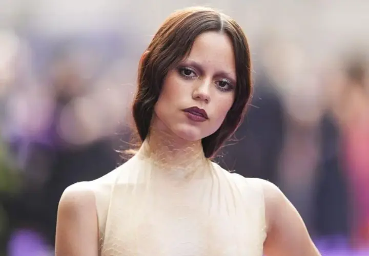 Jenna Ortega, czy ma polskie korzenie? Aktorka w kremowej sukni z siateczkowym kołnierzem, z burzą brązowych włosów i wyrazistym makijażem.
