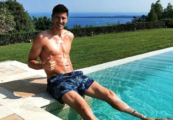 Robert Lewandowski w szortach z motywem liści siedzi na brzegu basenu, dając kciuk w górę. W tle błękitne morze i zieleń.
