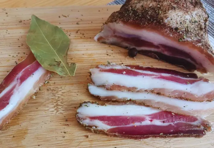 Pancetta co to? Pokrojony w plastry, przyprawiony boczek na desce. Obok liść laurowy i kawałek większego kawałka.