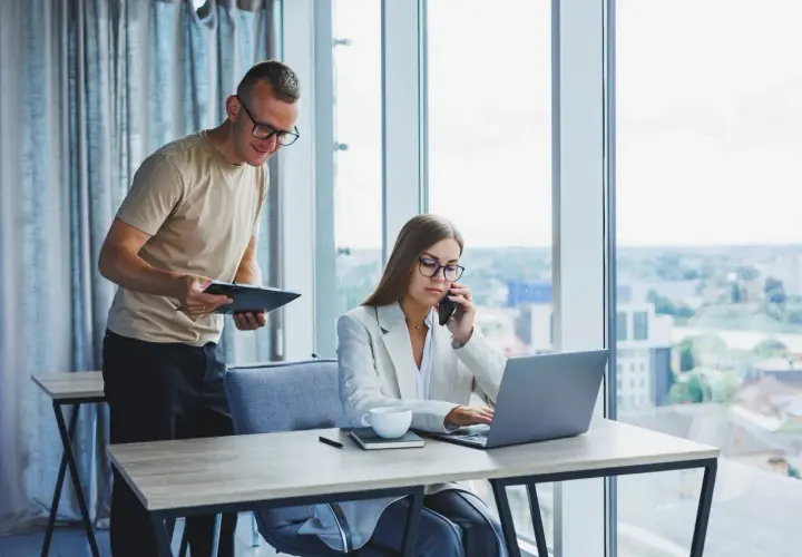 Un homme consulte un document pendant qu'une femme au téléphone travaille sur son ordinateur portable, illustrant l'efficacité de l'e-commerce B2B.