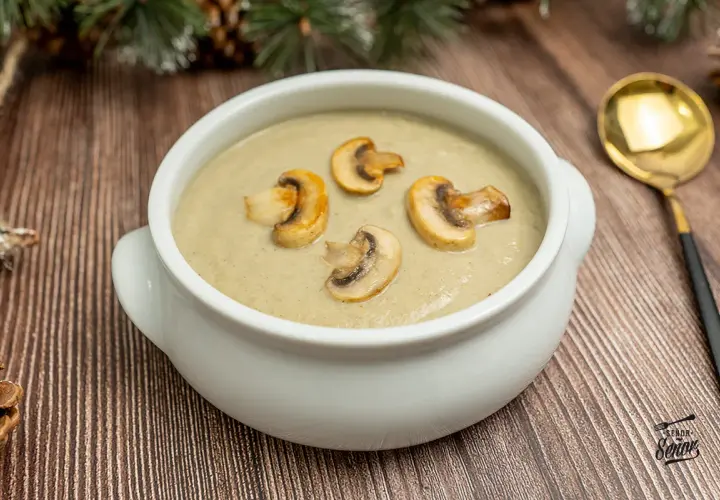 Crema de champiñones servida en un cuenco blanco, decorada con rodajas de champiñones salteados. Una cuchara dorada descansa a la derecha.