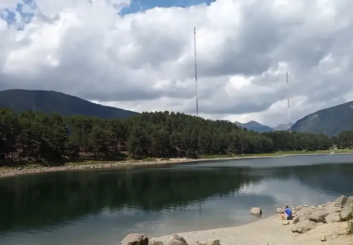 Persona sentada en la orilla del lago de Engolasters, rodeado de pinos y montañas, con dos torres de telecomunicaciones al fondo.