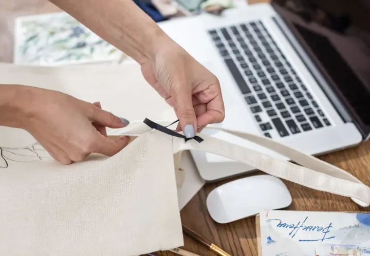 Ręce szyją torbę shopper, dodając czarną wstążkę. Na biurku laptop, myszka i materiały do szycia.