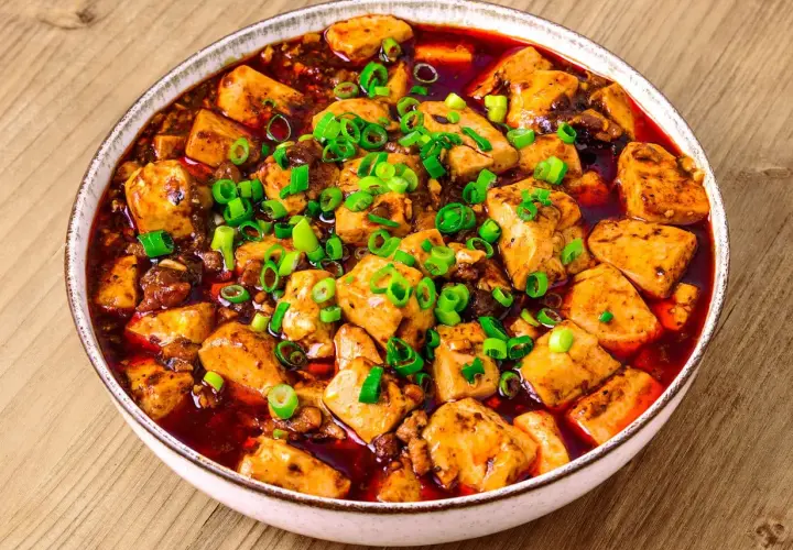 En skål med kryddig mapo tofu, toppad med färsk salladslök, serverad på ett träbord.