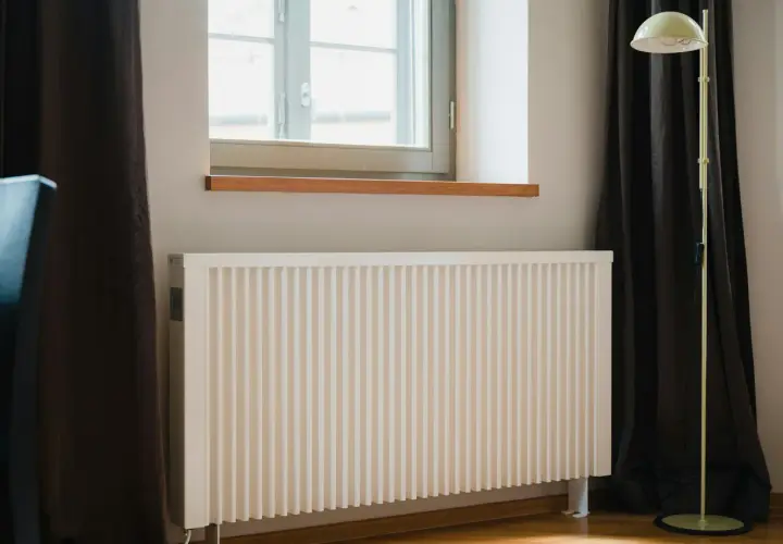 Vitt element med ribbad front, perfekt för att bygga elementskydd. Bredvid fönster med mörka gardiner och en golvlampa.