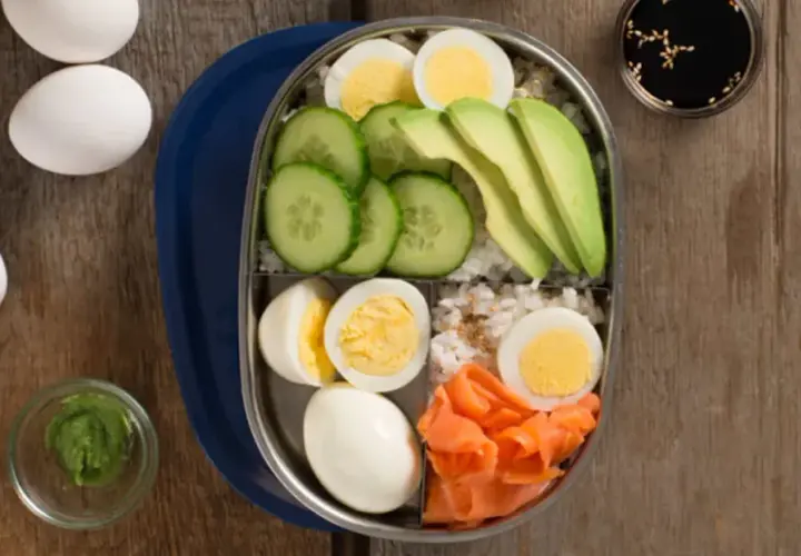 A bento box filled with rice, sliced cucumber, avocado, smoked salmon, and hard-boiled egg.