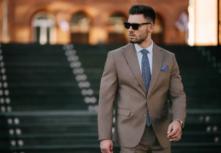 Elegancki mężczyzna w brązowej marynarce, z niebieską poszetką w kratkę i wzorzystym krawatem, w okularach przeciwsłonecznych.