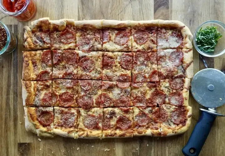 Prosty przepis na pizzę: prostokątna pizza pepperoni z serem, pokrojona na kwadraty, obok słoiczki z dodatkami i krajalnica.