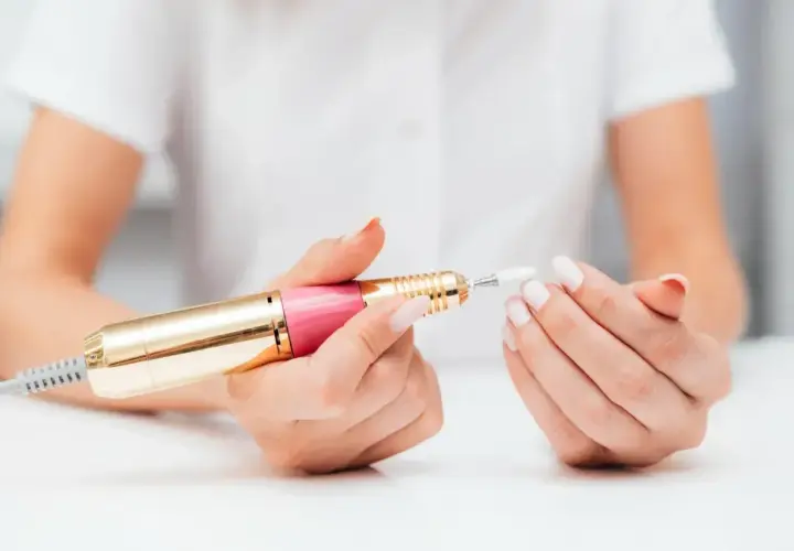 Dłonie z białymi paznokciami trzymają różowo-złotą frezarkę. Dowiedz się, jak używać frezarki do paznokci, aby uzyskać perfekcyjny manicure.