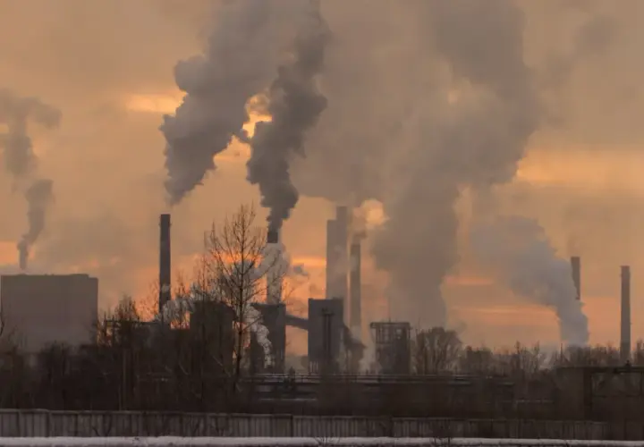 Chimeneas industriales expulsan humo denso, creando un paisaje sombrío. Las partículas contaminantes se elevan, tiñendo el cielo de un naranja opaco.