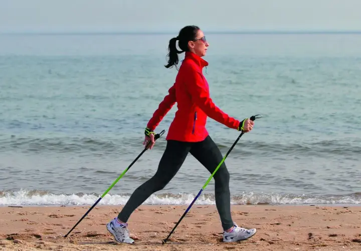 Kobieta w czerwonej kurtce ćwiczy marsz z kijkami na plaży. Pokazuje, jak chodzić z kijkami, wykorzystując je do aktywności fizycznej nad morzem.