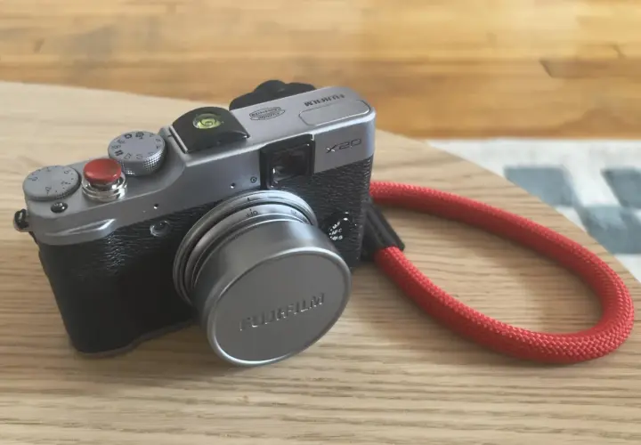 Srebrny aparat Fujifilm X20 z czerwoną smyczą leży na drewnianym blacie.