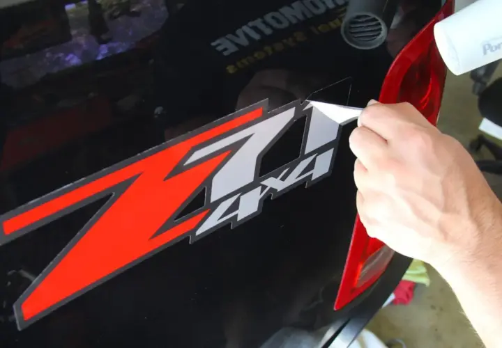 Mano che rimuove adesivo "Z7 4x4" da auto nera, usando un phon per ammorbidire il nastro adesivo vecchio.