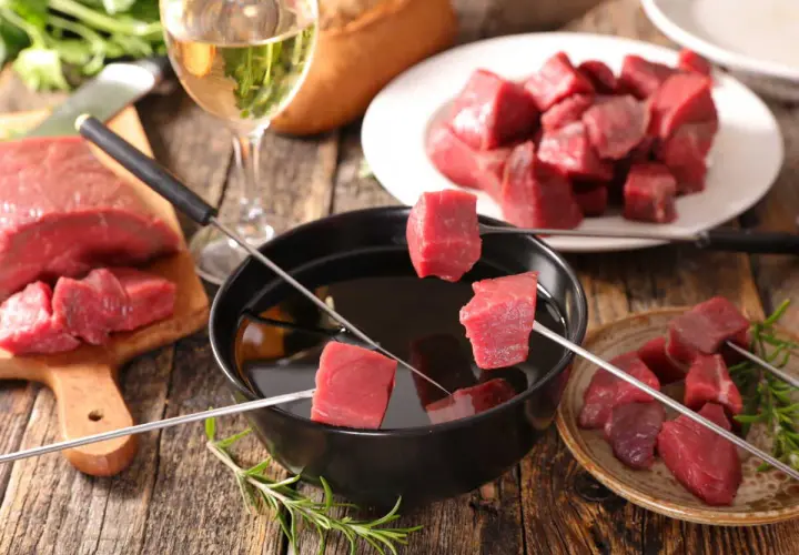 Préparer une fondue bourguignonne : des dés de bœuf crus sur des brochettes, prêts à être plongés dans l'huile chaude. Un verre de vin blanc à côté.
