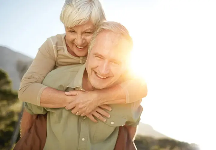 Ein Mann trägt eine Frau auf dem Rücken. Beide lachen und genießen die Sonne. Ein Bild von wahrer **Liebe im Alter**.