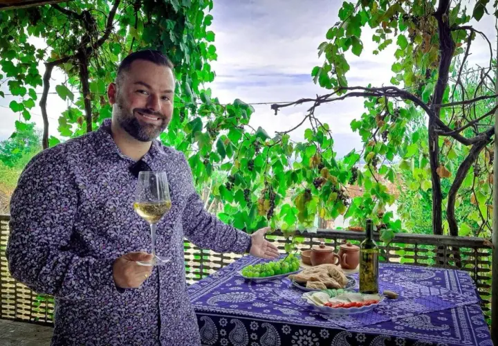 Mężczyzna z kieliszkiem białego wina, pod winoroślą, zaprasza do degustacji marani wino. Na stole owoce, chleb i ser.