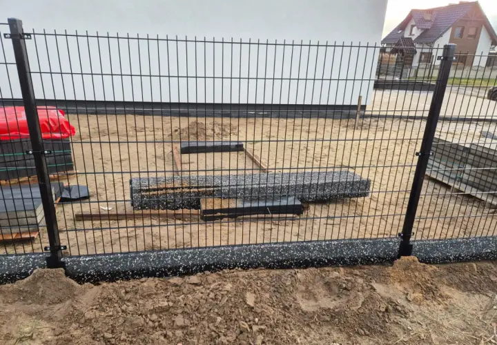 Elementy ogrodzenia panelowego w trakcie montażu. Widać słupki, panele i materiały budowlane na placu budowy.