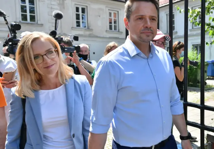 Małgorzata Trzaskowska w jasnej marynarce i Rafał Trzaskowski w błękitnej koszuli otoczeni przez dziennikarzy.