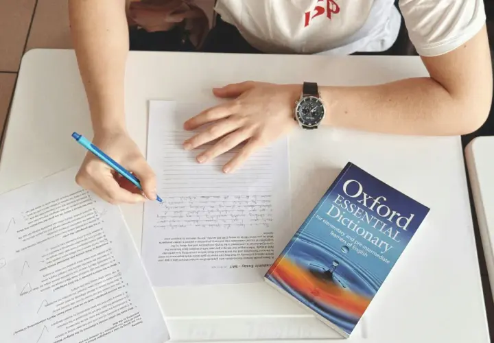 Studente che scrive, con un dizionario Oxford Essential Dictionary aperto. Cosa vuol dire AT? Forse "at" è un'abbreviazione usata in questo contesto di studio.