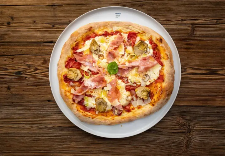 Eine köstliche Pizza mit Prosciutto Crudo, Artischocken und frischem Basilikum auf einem weißen Teller, serviert auf rustikalem Holz.