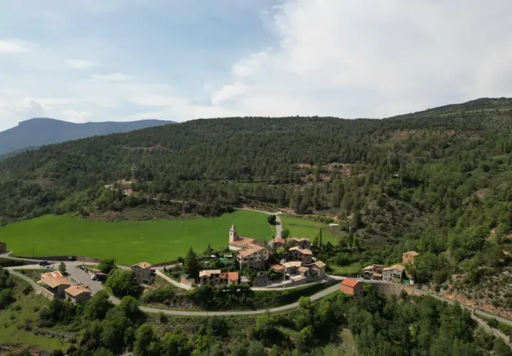 Vista aérea del pintoresco pueblo de **Vall de la Vansa**, rodeado de verdes prados y frondosos bosques, con casas de piedra y una iglesia.