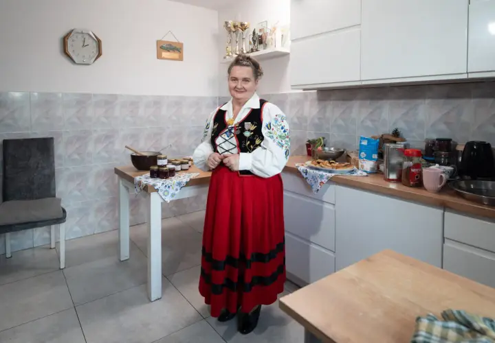 Kobieta w stroju ludowym stoi w kuchni obok stołu z dżemami. Na ścianie wisi zegar, wskazujący godzinę 10.