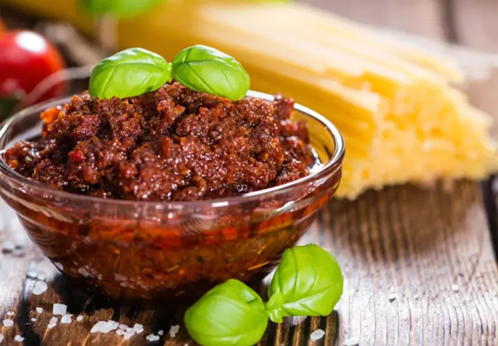 Pesto di pomodori secchi in una ciotola di vetro, con spaghetti e basilico sullo sfondo.