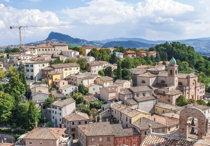 Panoramica di un borgo collinare con case dai tetti rossi, una chiesa con campanile e montagne sullo sfondo. Un paesaggio incantevole nei dintorni di Rimini.