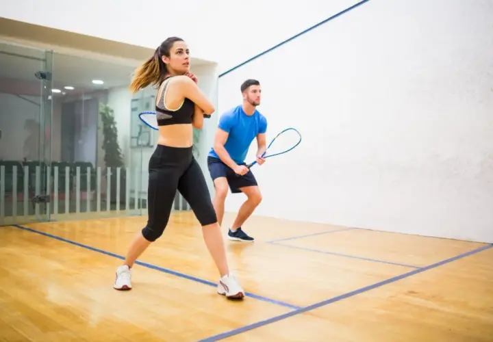 Para ćwiczy squasha, poznając zasady gry. Kobieta w sportowym stroju i mężczyzna w niebieskiej koszulce z rakietami gotowi do akcji na boisku.