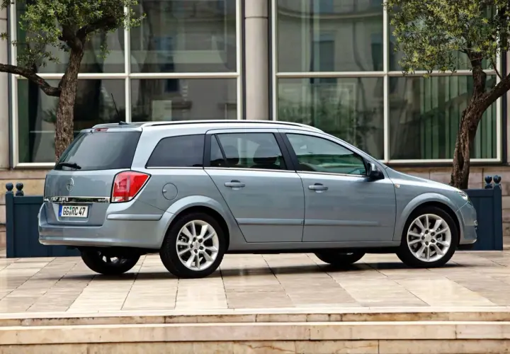 Srebrny Opel Astra 2005 kombi zaparkowany przed nowoczesnym budynkiem z drzewami.