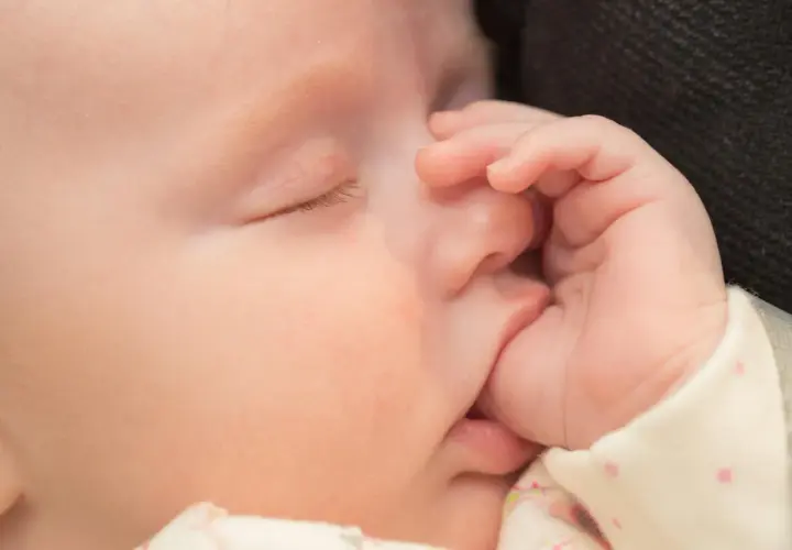Uroczy niemowlak śpi, a jego mała rączka delikatnie obejmuje usta. To naturalne ssanie kciuka u niemowlaka, które przynosi mu ukojenie.