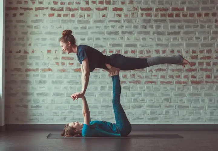 Dwie kobiety ćwiczą **acroyoga** na macie. Jedna leży na plecach, podtrzymując drugą, która unosi nogi w powietrzu.