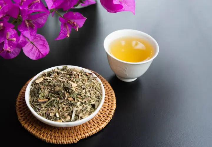 Una tazza di tisana per digerire accanto a una ciotola di erbe secche, con fiori viola sullo sfondo.