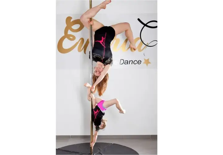 Dwie dziewczyny ćwiczą pole dance. Młodsza wspiera starszą, która wisi głową w dół.