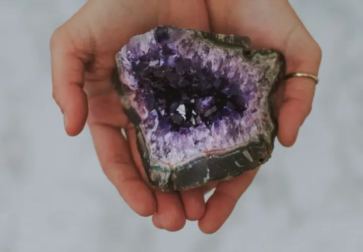 Hände halten einen tiefvioletten Amethyst. Seine Wirkung auf die Haut wird oft als beruhigend beschrieben.