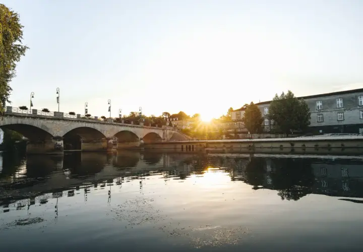 Un pont de pierre traverse une rivière calme, reflétant le soleil couchant sur les bâtiments historiques. Une belle vue pour savoir quoi faire à Cognac.
