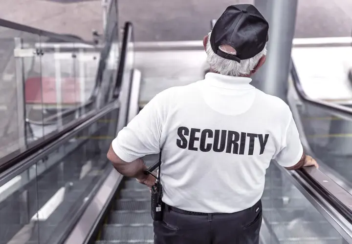 Doświadczony technik ochrony fizycznej osób i mienia, w białej koszulce z napisem "SECURITY", stoi na ruchomych schodach.