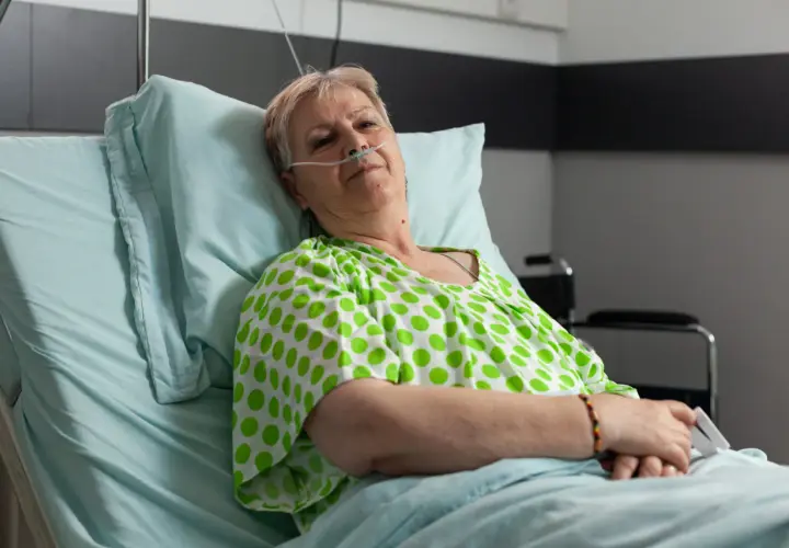Starsza kobieta w szpitalnym łóżku, z tlenem przy nosie, w zielono-białej bluzce w groszki. Czeka na leki, być może Duracef.
