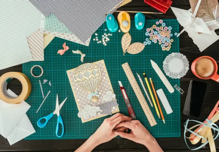 Ręce tworzą kartkę z balonem na gorące powietrze. Wokół materiały do scrapbookingu, taśma, nożyczki, guziki i inne akcesoria.