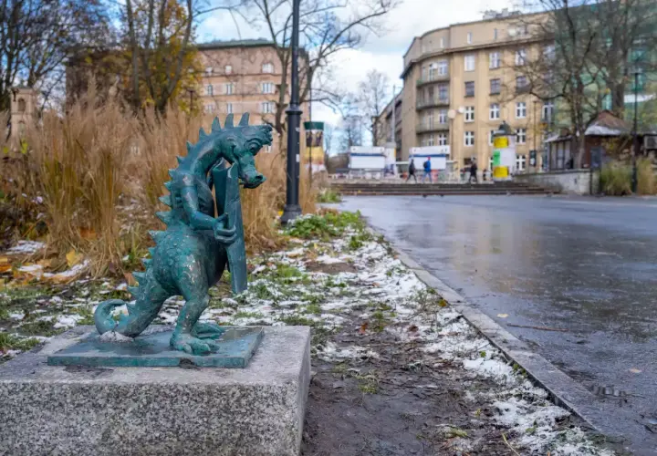 Mały smok z brązu, symbol smoczy szlak kraków, stoi na kamiennym postumencie. W tle miejski krajobraz z budynkami i mokrą nawierzchnią.