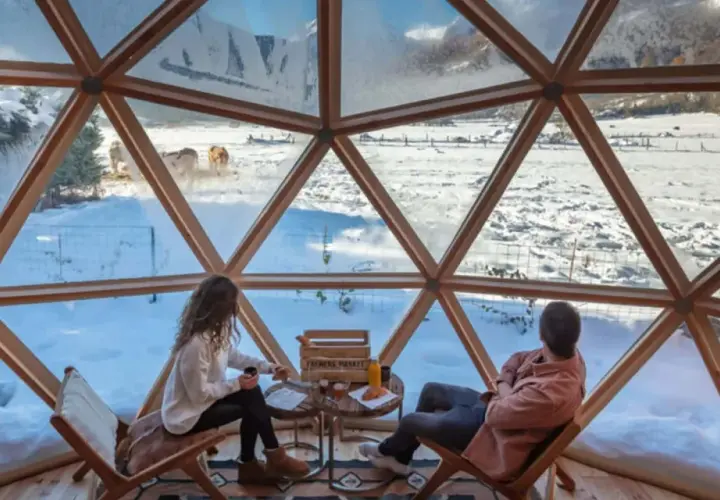 Pareja disfruta de un paisaje nevado en el Pirineo francés desde una cúpula geodésica. Vacas pastan en la distancia.