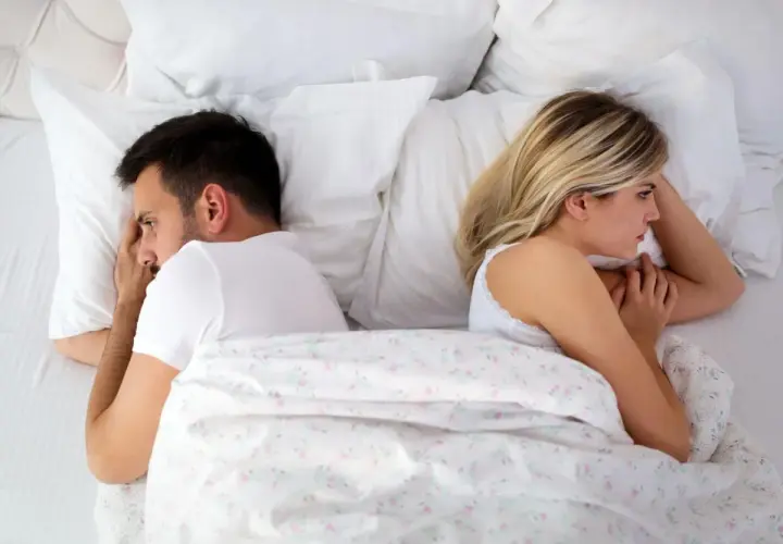 Coppia a letto, distanti. Lei, con lo sguardo perso, sembra non avere voglia di fare l'amore. Lui, pensieroso, si volta dall'altra parte.