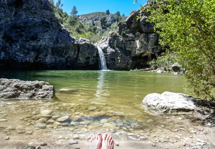 Pies descalzos en la orilla del **barranco de la Encantada**, con una cascada cayendo en una poza de agua verde rodeada de rocas y vegetación.