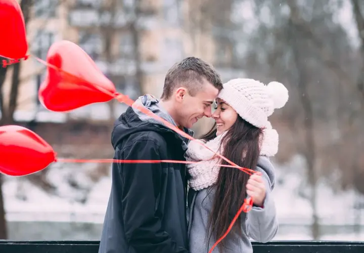 Para wzruszona, trzymająca czerwone balony w kształcie serca. Chłopak szepcze jej do ucha, co napisać dziewczynie żeby się wzruszyła.