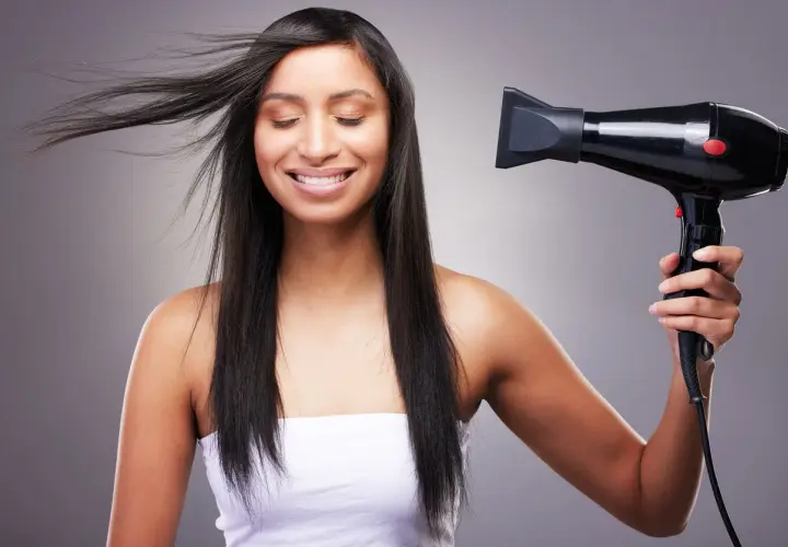 Mujer sonríe mientras se seca el pelo fino y escaso con un secador negro.