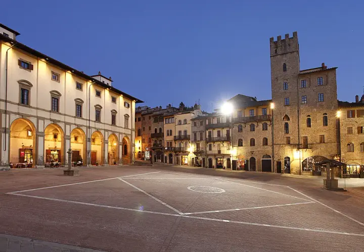 Piazza illuminata ad Arezzo, con portici, edifici storici e una torre. Sembra proprio che la vita è bella ad Arezzo.