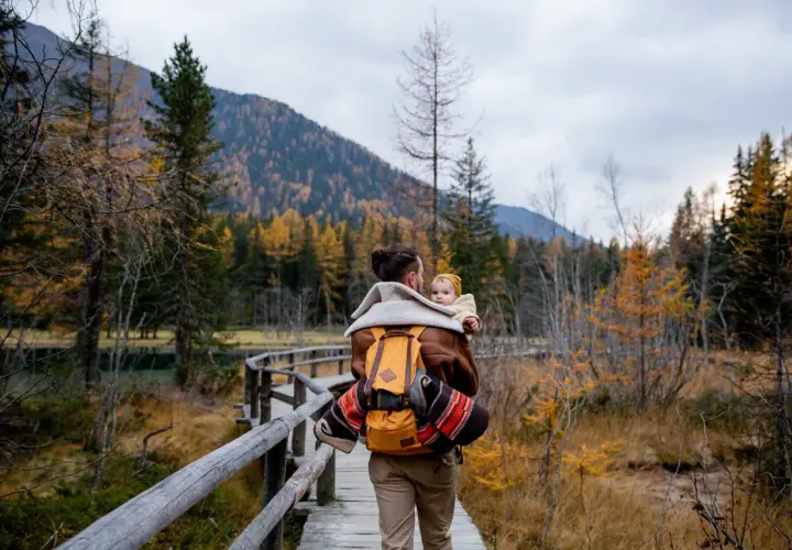 Ojciec z dzieckiem na plecach spaceruje drewnianą kładką pośród jesiennych lasów. Tatry z dzieckiem to piękna przygoda.
