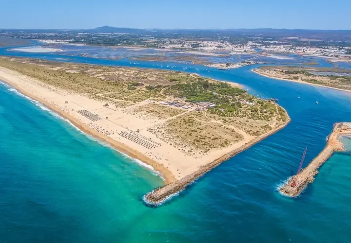 Vista aérea de la **isla de Tavira**, con su extensa playa de arena dorada, aguas turquesas y el canal que la separa de tierra firme, donde se aprecian salinas y la ciudad al fondo.